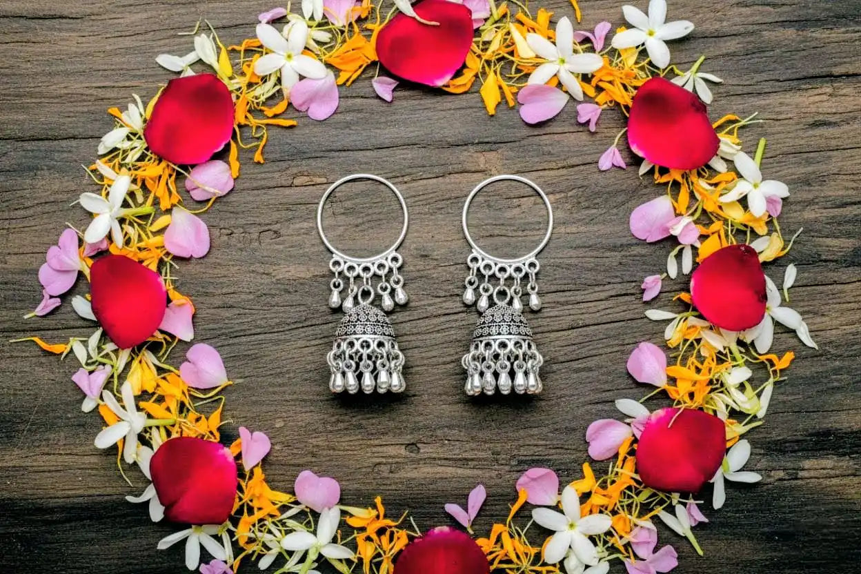 Mayuri Jhumka – oxidized silver hoop jhumka earrings arranged inside a floral ring of rose, jasmine, and marigold petals on a rustic wooden background from Shinora