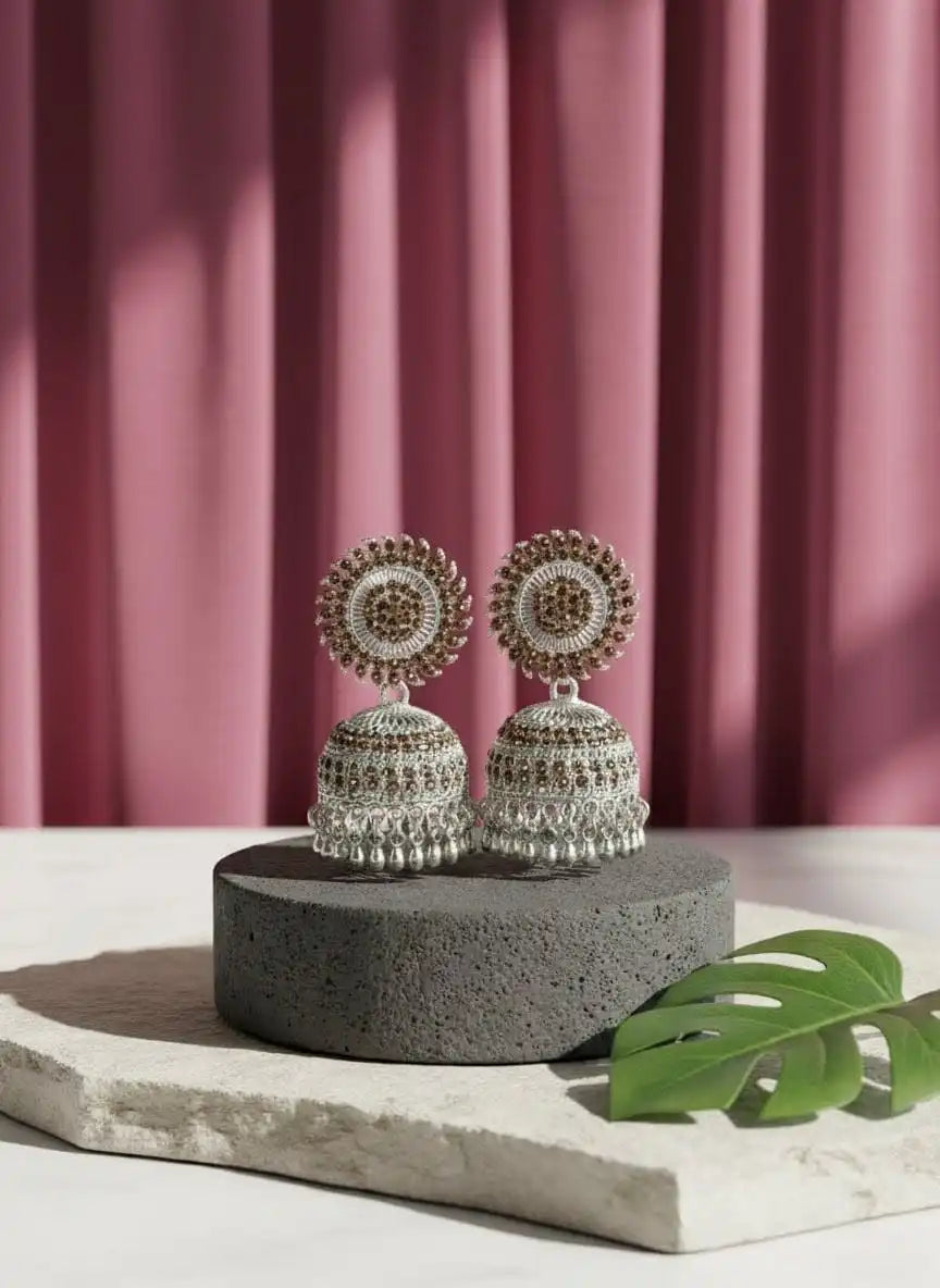 Dia Jhumka – intricately crafted gold-tone jhumka earrings with brown stones and ghungroo fringe displayed on a textured stone platform with a pink backdrop from Shinora