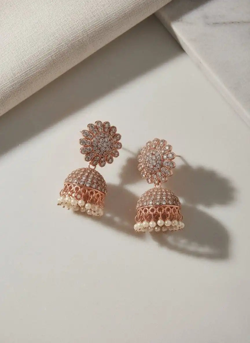 Noor Jhumka – sparkling rose-gold jhumka earrings with stone-studded floral tops and pearl drops on a soft beige backdrop from Shinora