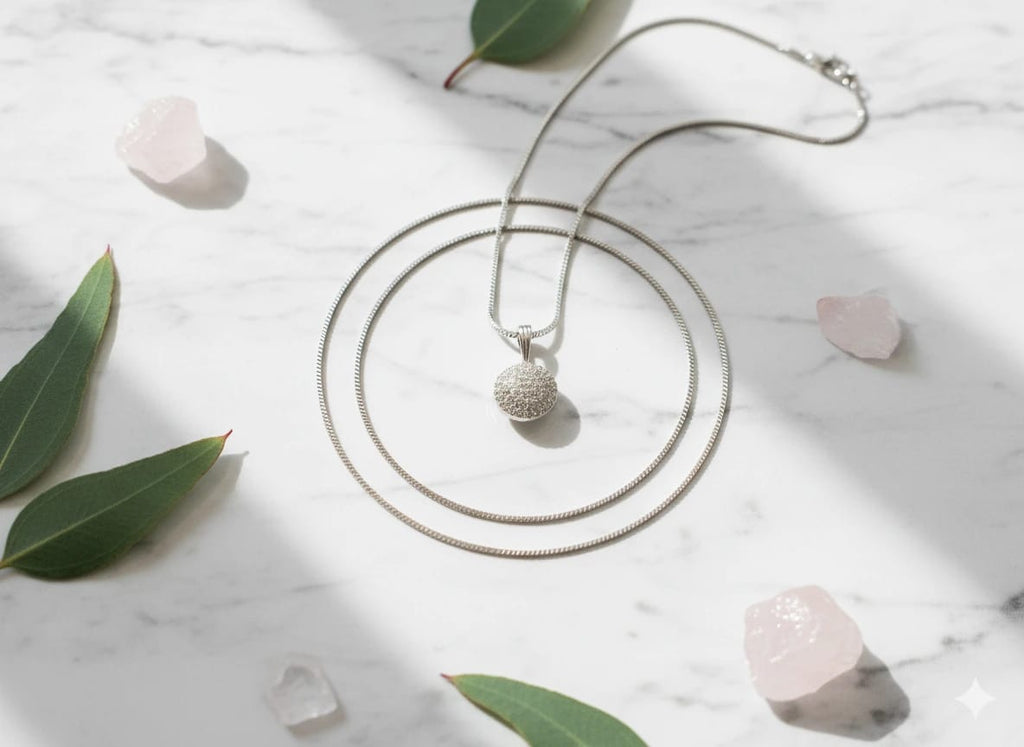 Elegant silver round pendant necklace with crystal detailing, displayed on a white marble surface with green leaves and rose quartz stones – The Celeste Necklace by The Fluttering Charm by Shnora