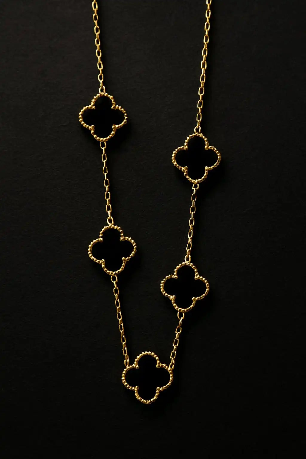 Gold clover necklace hanging on textured black background highlighting shiny finish and clover detailing from shinora